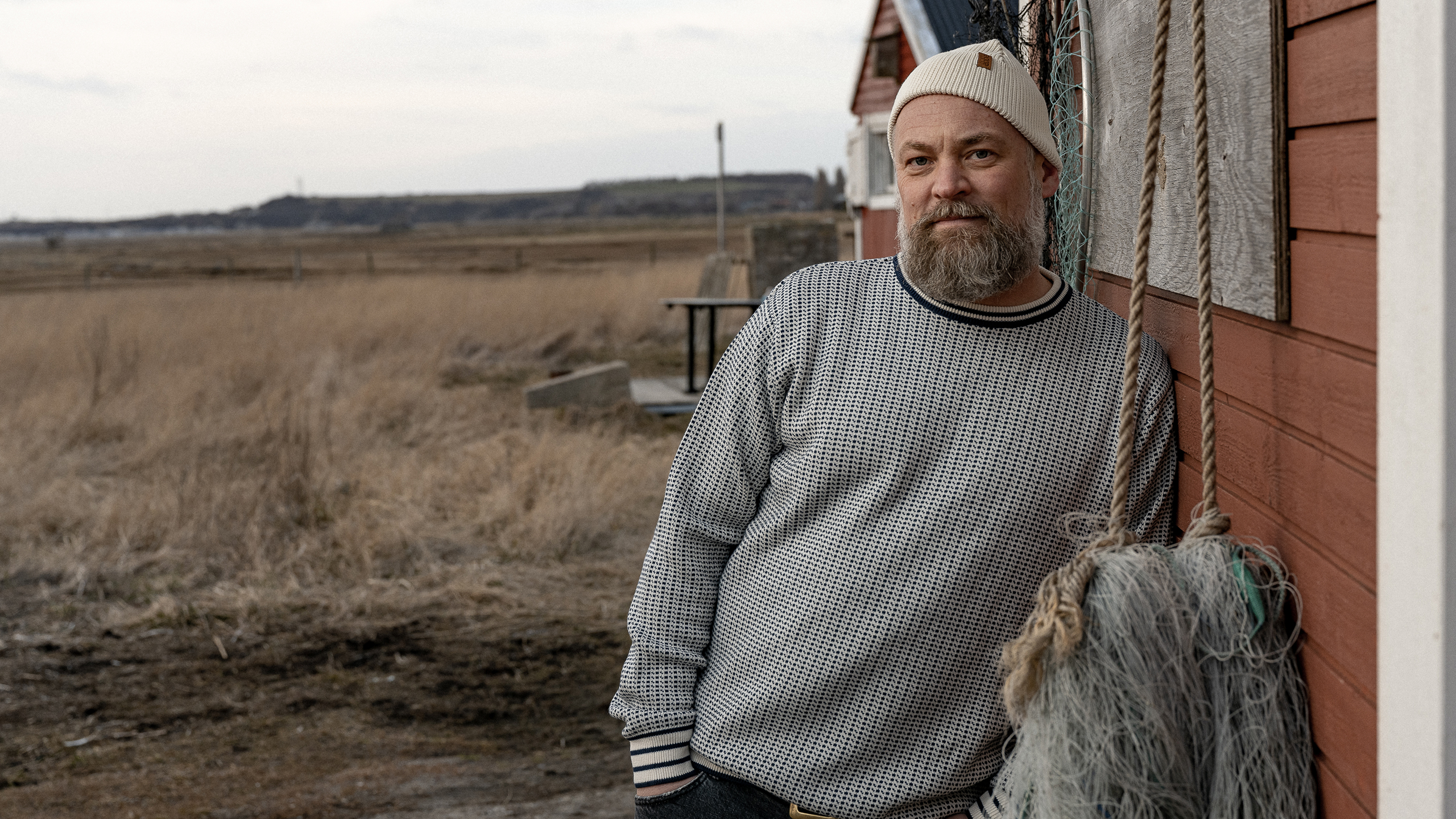 Man leaning on a wall. Soft evening light. Man has a big beard and a knitted sweater.